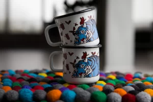 Two white enamel mugs with blue and red illustrations of tigers and butterflies, stacked on top of each other. The upper mug is slightly off focus, with the brand logo 'emalco enamelware' visible in the upper left corner. The mugs are placed on a surface with a colorful, fuzzy background consisting of what appears to be a multicolored felt ball mat.